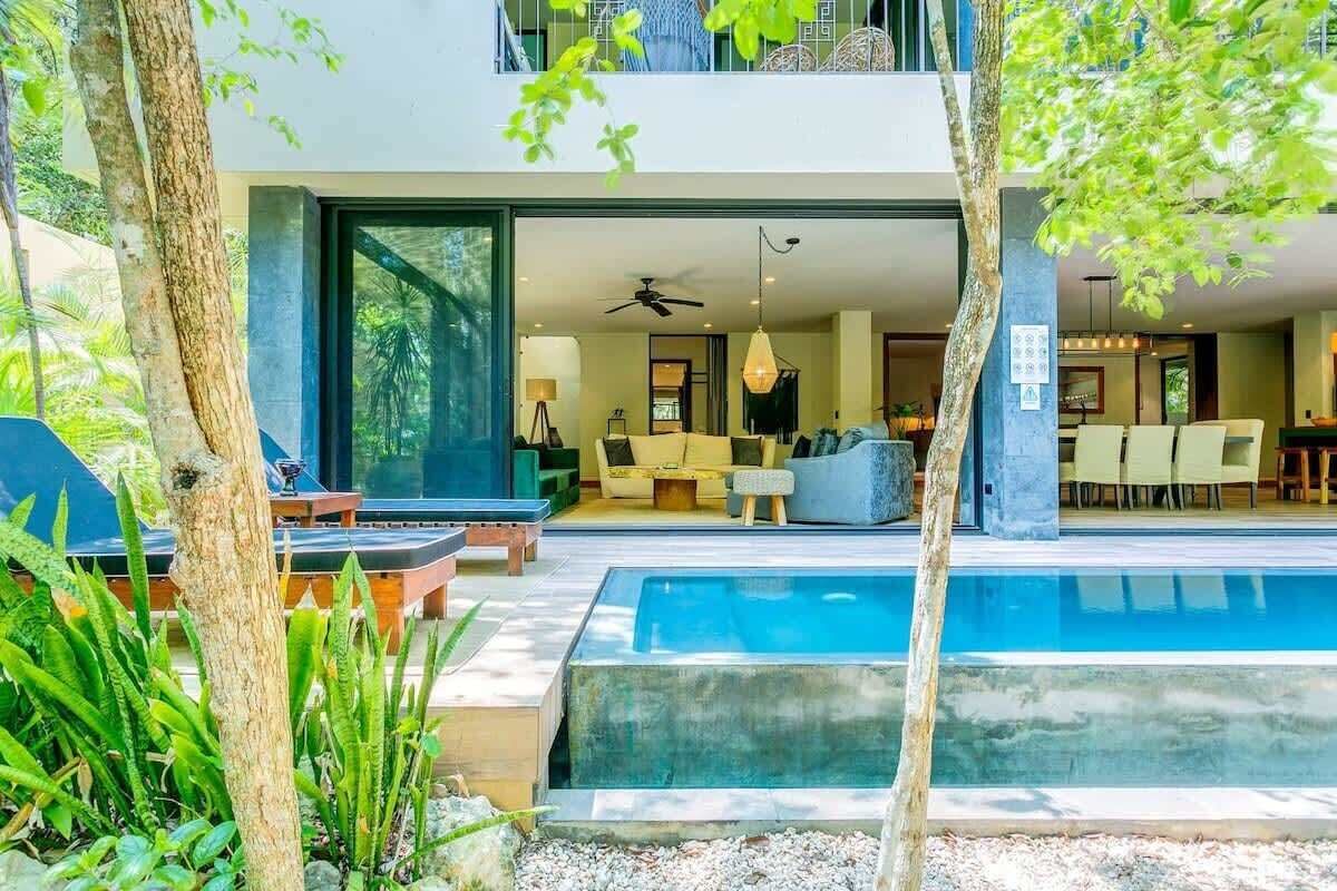 Un área de piscina moderna al aire libre, con un espacio para comer y una sala de estar visible a través de un ventanal abierto.