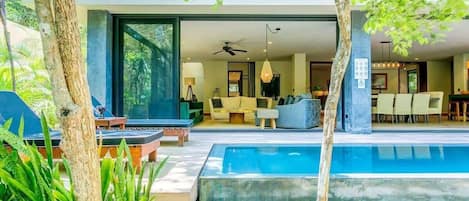 Un área de piscina moderna al aire libre, con un espacio para comer y una sala de estar visible a través de un ventanal abierto.
