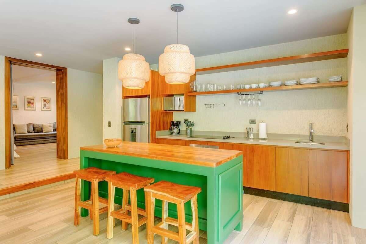 A modern kitchen with a green island, wooden stools, and pendant lighting.