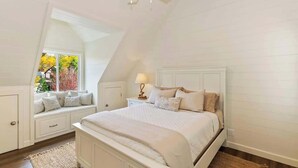 Un dormitorio con techo inclinado, una cama con ropa de cama blanca, una mesita de noche con una lámpara y una ventana con vistas a árboles.