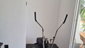 Un vélo d’appartement est installé dans une pièce dont le sol est noir, un mur est blanc et une porte-fenêtre coulissante donne sur un espace extérieur où se trouvent une chaise en osier et un pot de fleurs.