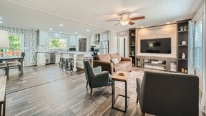 A spacious living area with a sofa, chairs, a coffee table, a TV, and a kitchen in the background. - Secluded Country Home on 15 Acres with a BBQ and Upscale Kitchen. Close to COTA and the Airport. (Cedar Creek)