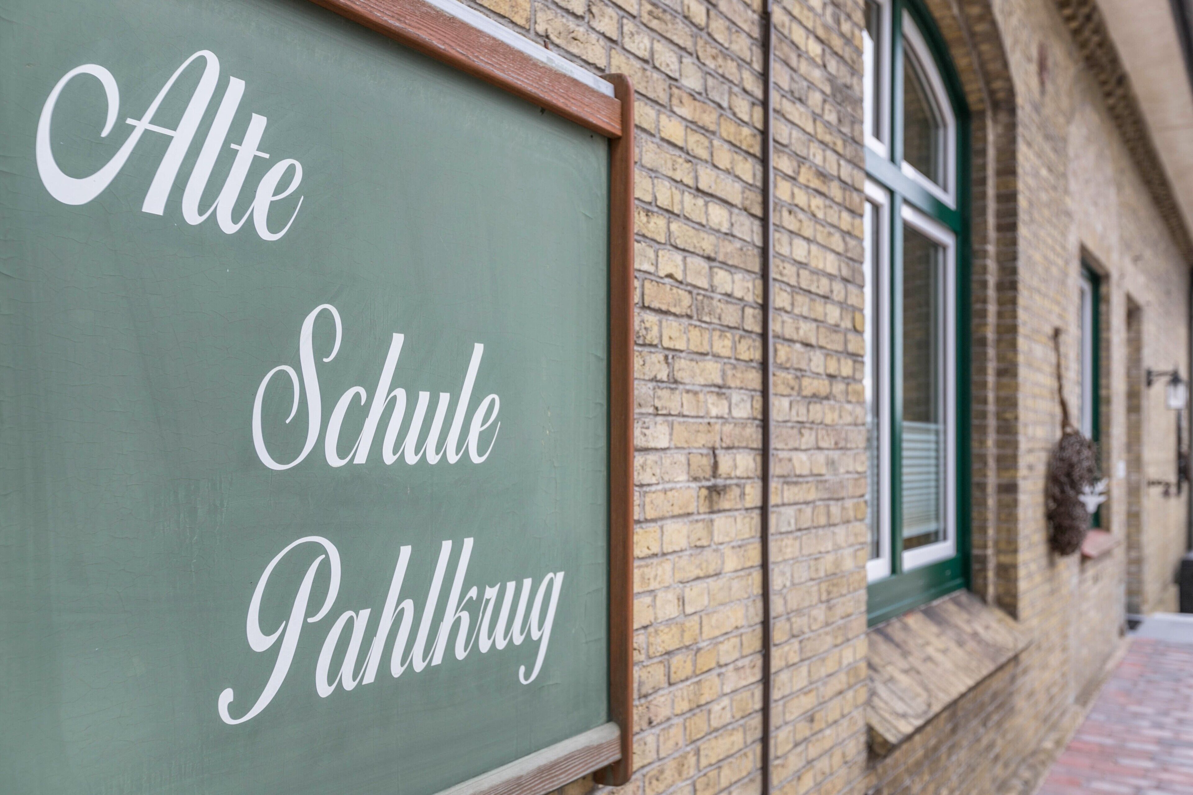 En tavle med 'Alte Schule Pahlkru' skrevet på, fastgjort til en murbygning med et vindue.