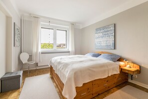 A bedroom with a wooden bed, a side table with a lamp, a chair, and a window with curtains. - Apartment 'Alte Schule Pahlkrug' with Private Terrace, Private Garden and Wi-Fi (Linden)