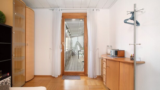 A compact room with a wooden wardrobe, a small cabinet, a coat rack, and a view through a doorway.