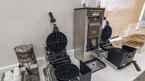 A waffle station with two waffle makers, a syrup dispenser, and a container of waffles. - MainStay Suites Chambersburg (Chambersburg)
