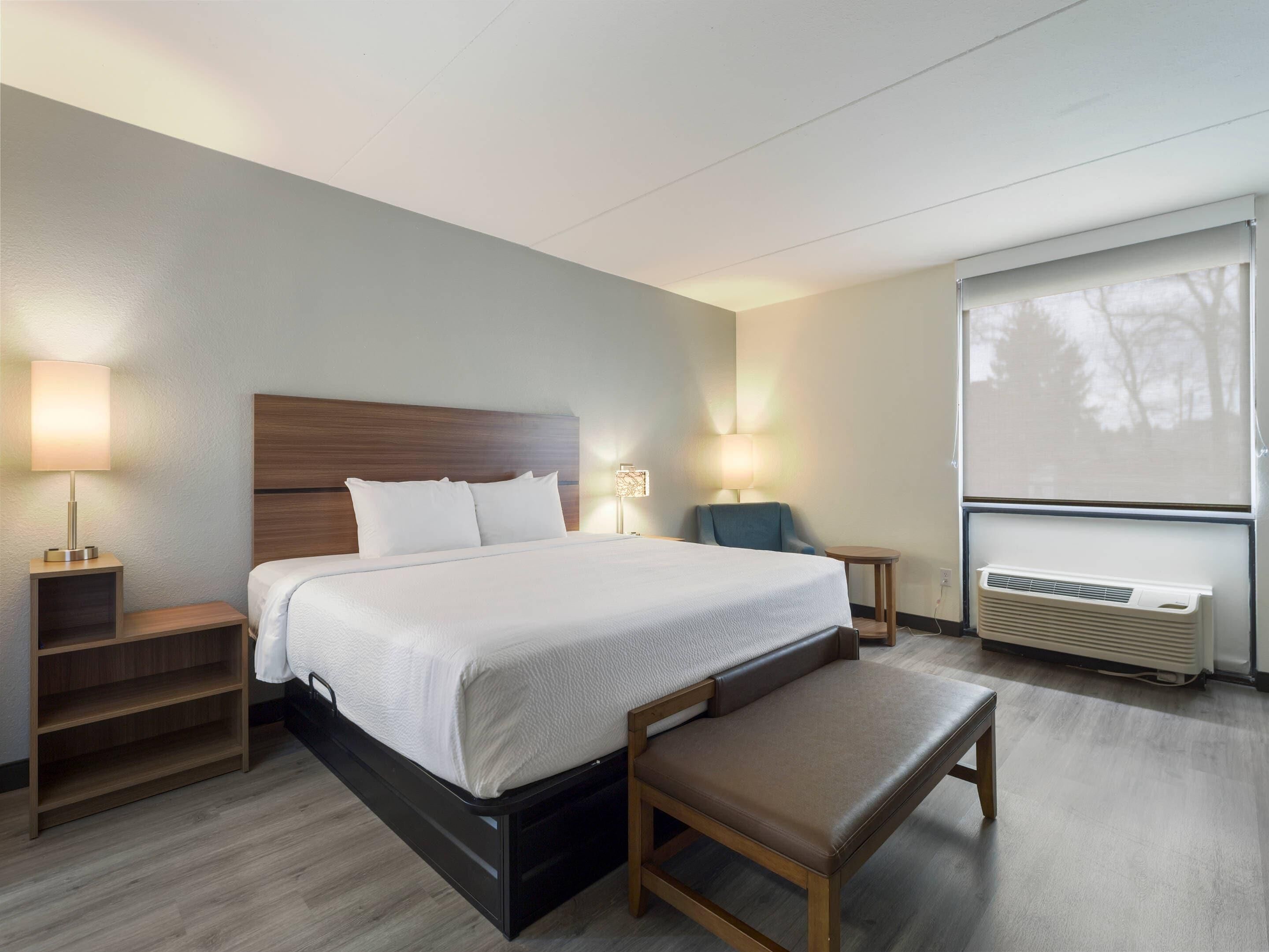A hotel room with a bed, bedside tables, a bench, a chair, a standing lamp, a desk lamp, and an air conditioning unit.