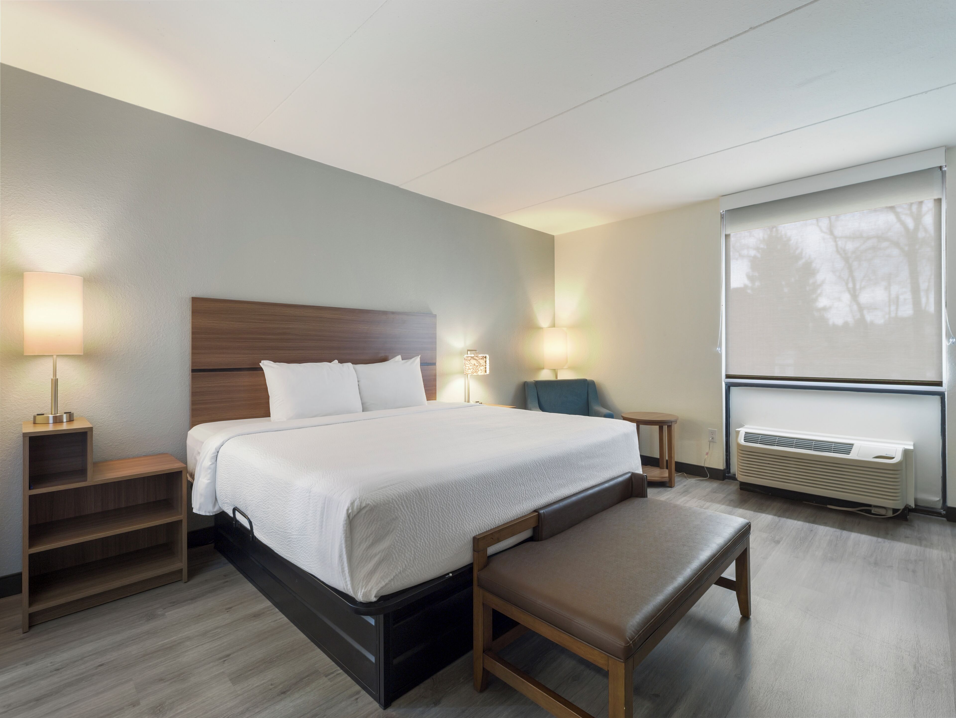 Ein Hotelzimmer mit Bett, Nachttischen, einer Bank, einem Sessel, einer Wand-Klimaanlage und einem Fenster mit Blick auf Bäume.