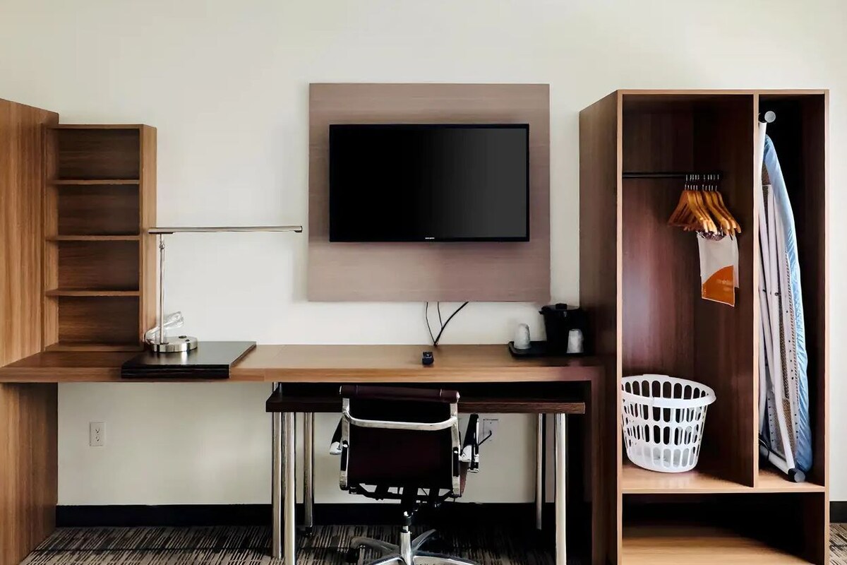 A modern hotel room with a wooden desk, a mounted flat-screen TV, a chair, a wardrobe with hanging clothes, and a laundry basket.