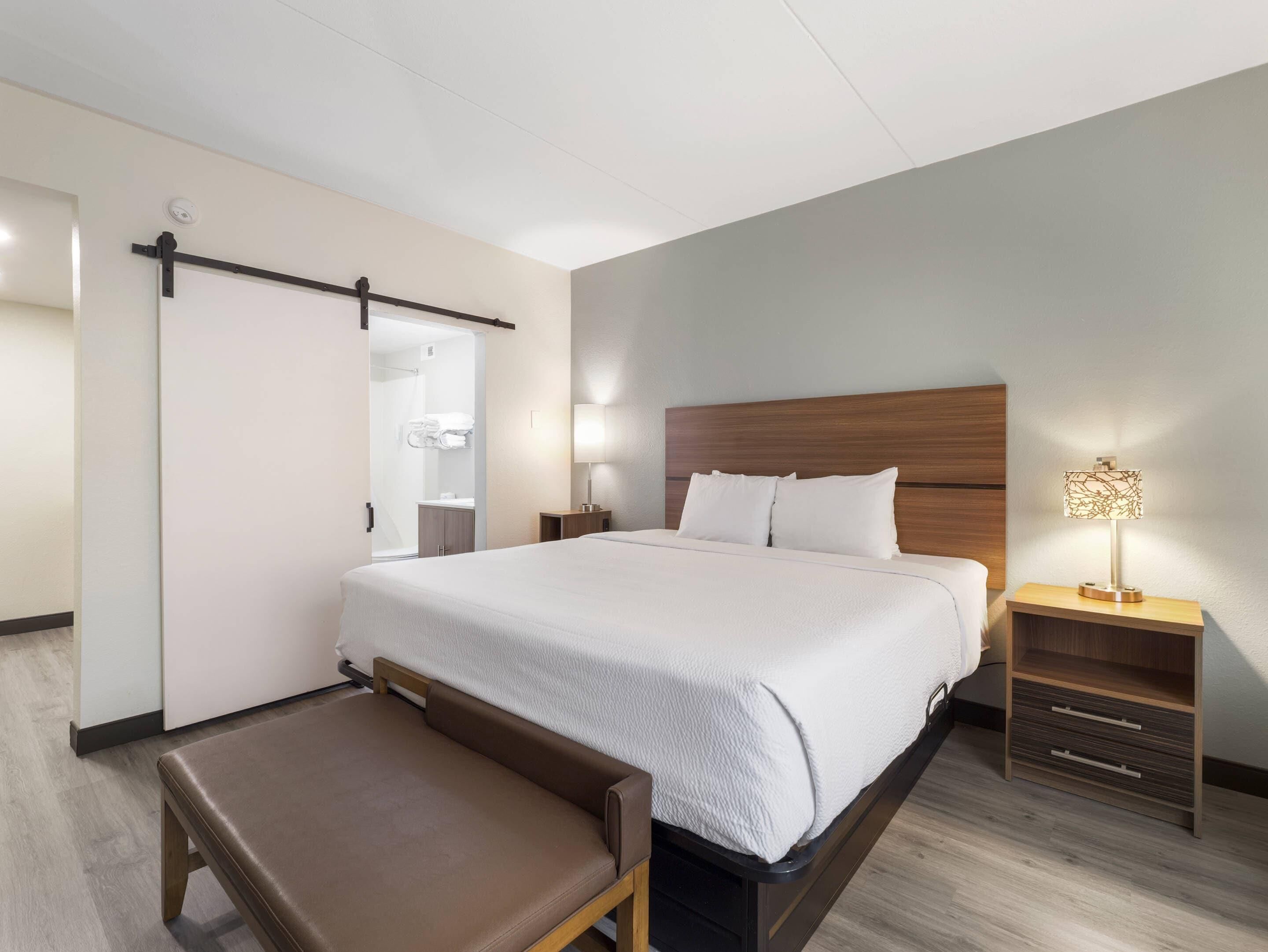 A modern hotel room with a bed, bedside table, lamp, and a bench.