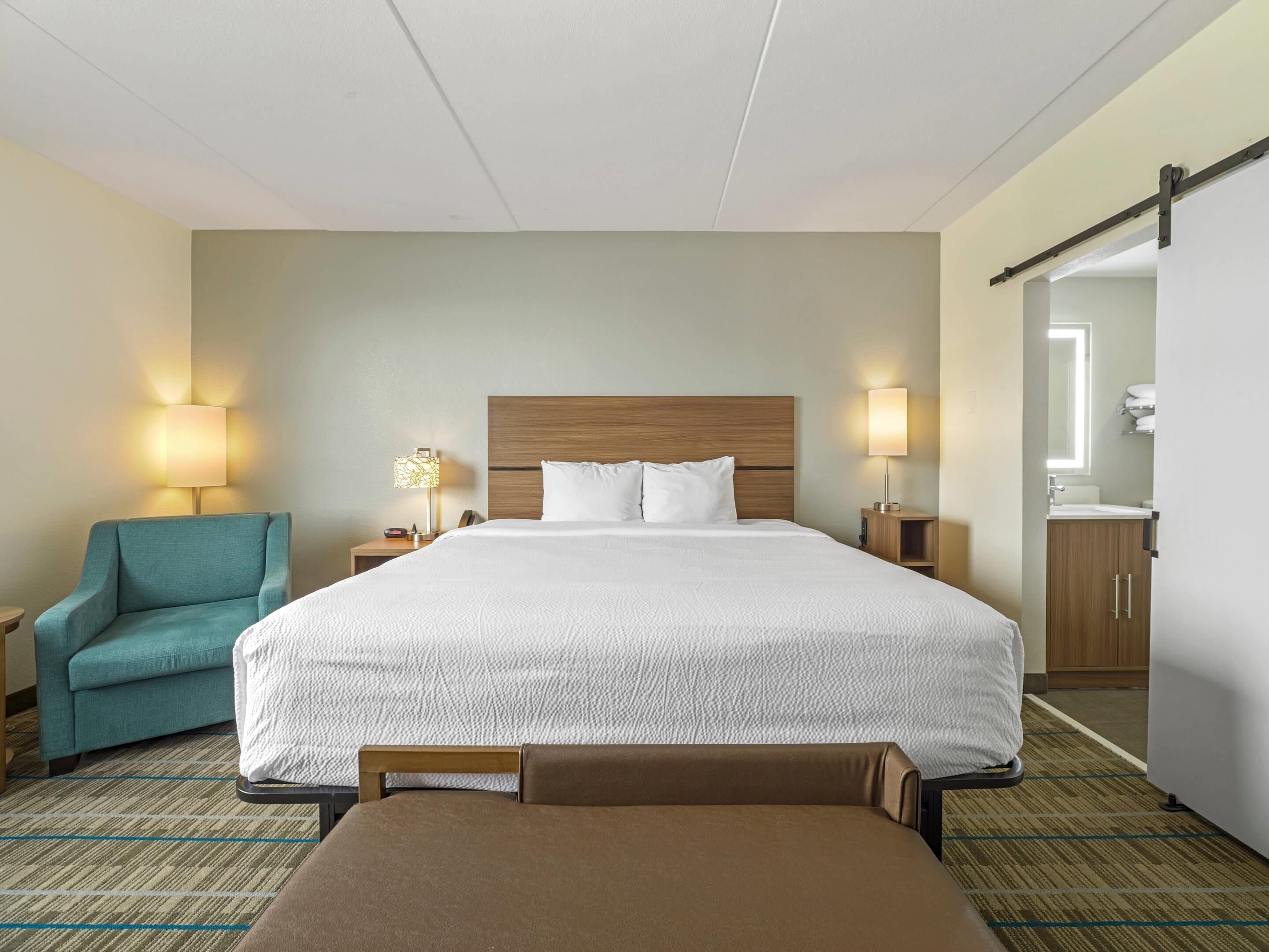 Une chambre d’hôtel avec un grand lit, deux tables de chevet avec des lampes, une chaise, un banc et une salle de bain avec un miroir et un lavabo.