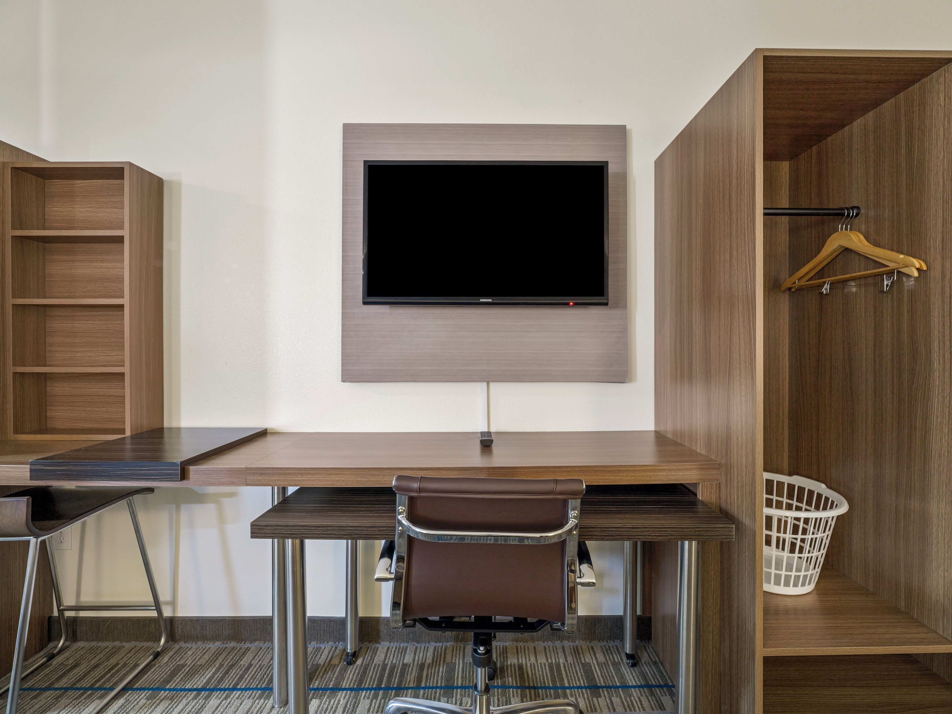 Ein modernes Zimmer mit Flachbildfernseher an der Wand, einem Schreibtisch mit Stuhl und einem Holzschrank mit Kleiderstange.
