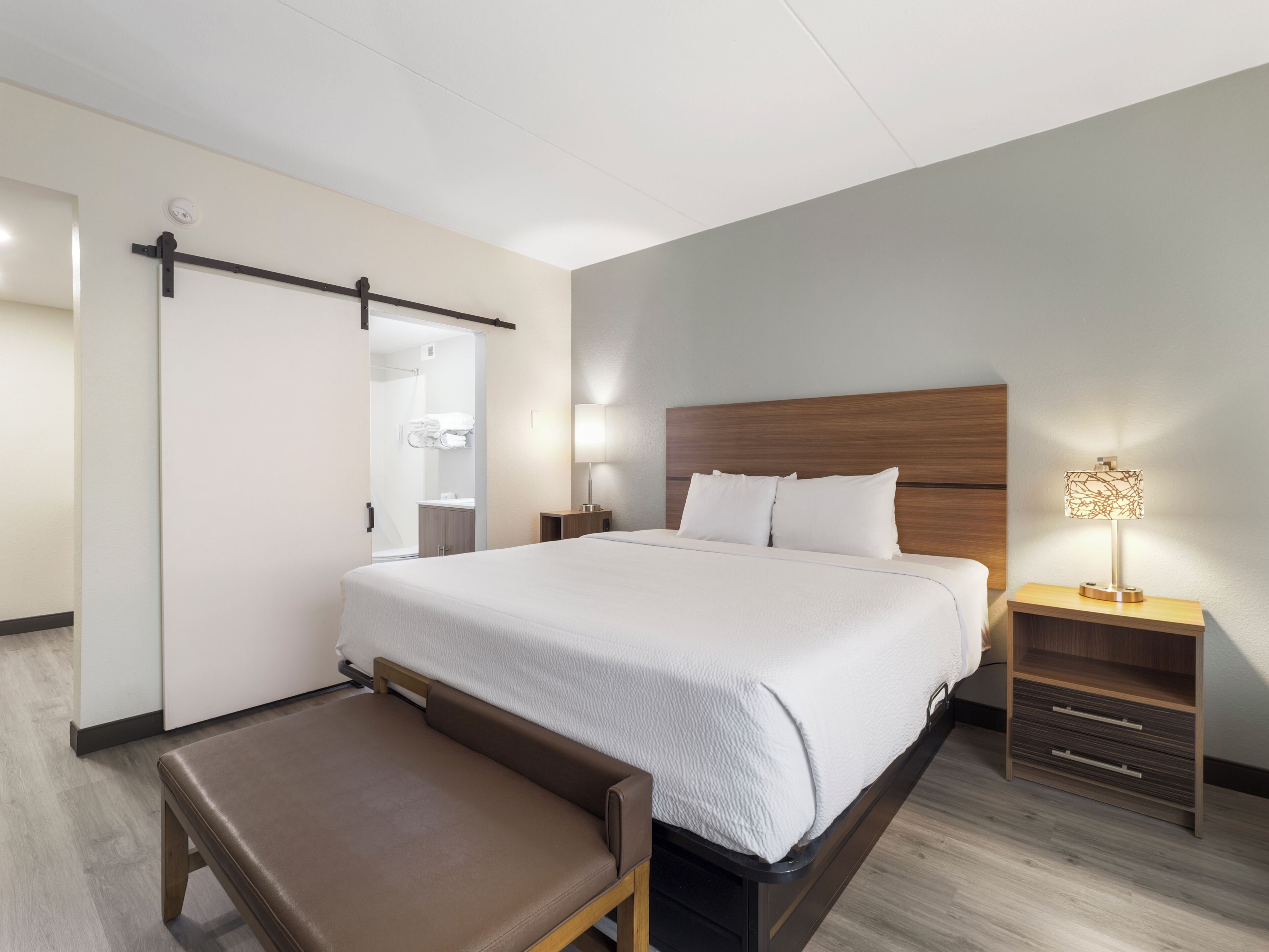 Habitació d'hotel moderna amb un llit gran, una banca, una taula de nit amb una làmpada i un armari encastat.