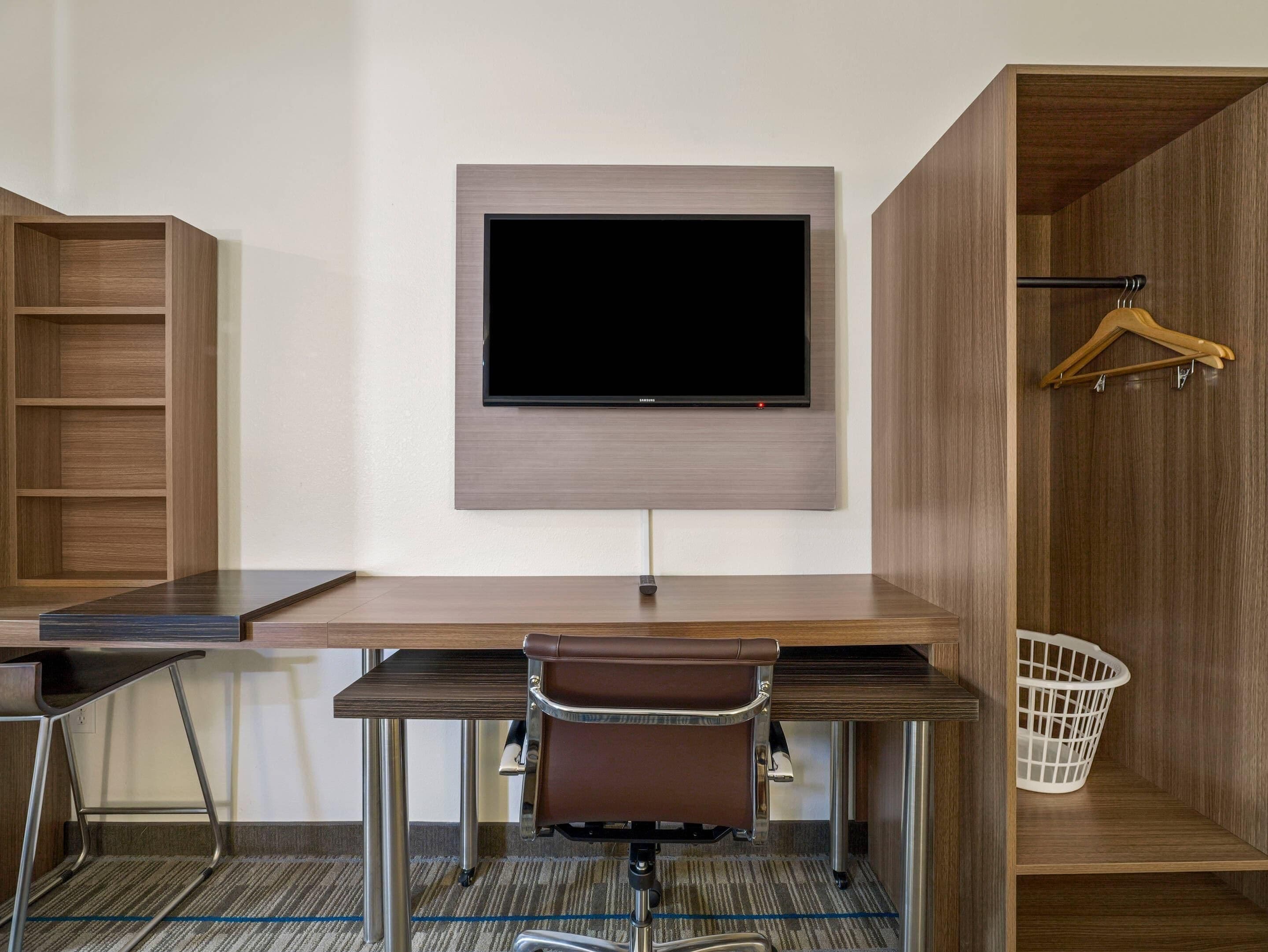 A modern hotel room with a flat-screen TV mounted on the wall, a desk with a chair, a coat rack, and a laundry basket.
