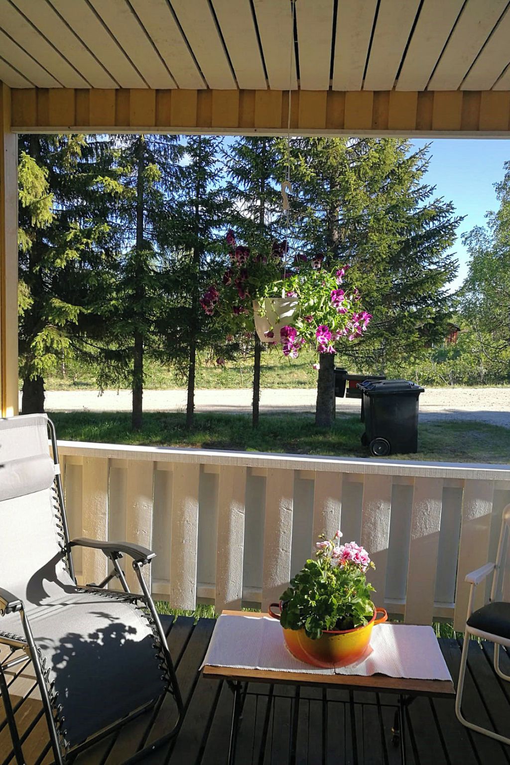 A covered outdoor seating area with a table and chairs, surrounded by trees and a trash bin.