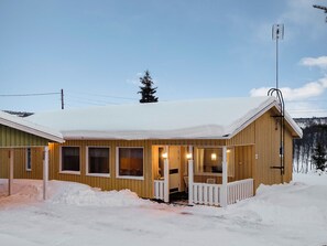 Una cabaña de madera con un porche, varios ventanales y un paisaje cubierto de nieve.