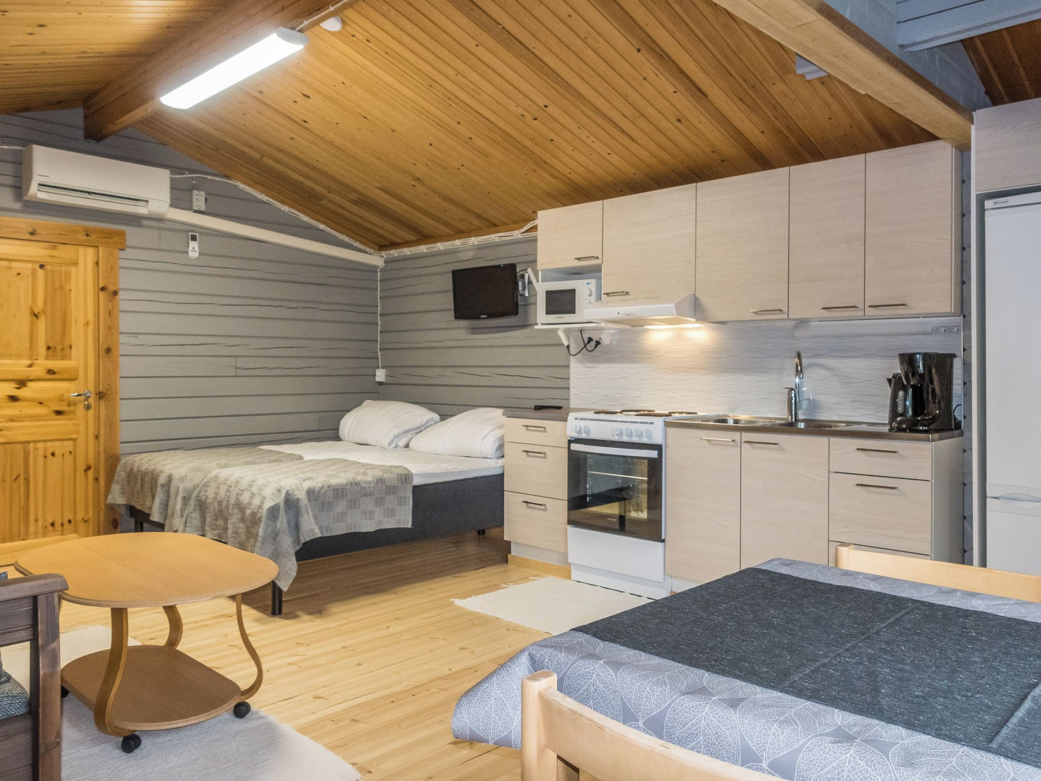 A compact room with a bed, a small table, a kitchenette, and a wooden ceiling.