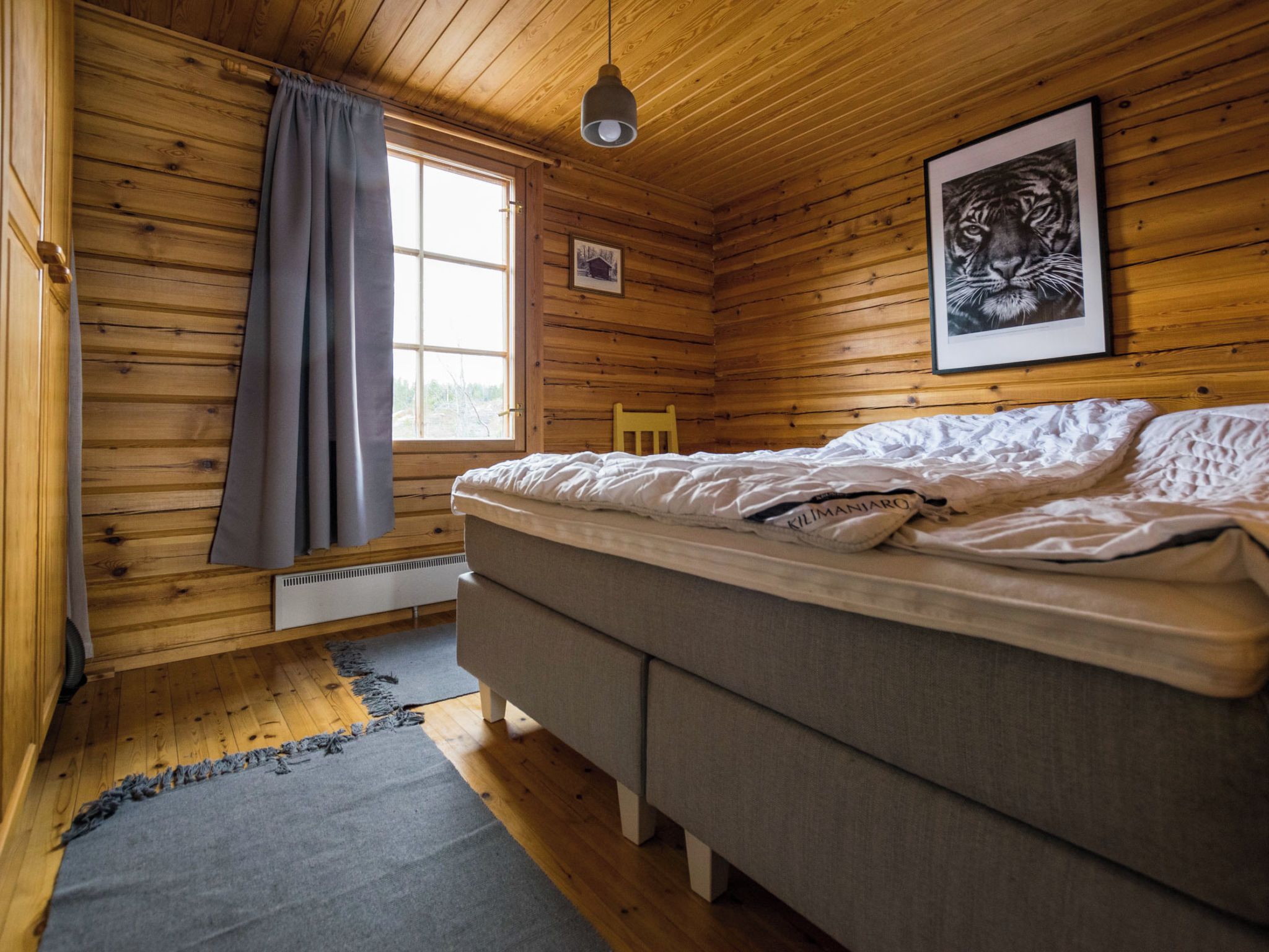 A wooden cabin room with a bed, a window with curtains, a framed picture on the wall, and a hanging light fixture.