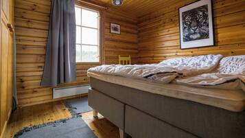 A wooden cabin room with a bed, a window with curtains, a framed picture on the wall, and a hanging light fixture.