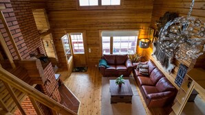 Een gezellig houten blokhutinterieur met een slaapkamer op een entresol, houten wanden, een open haard en een groot raam.
