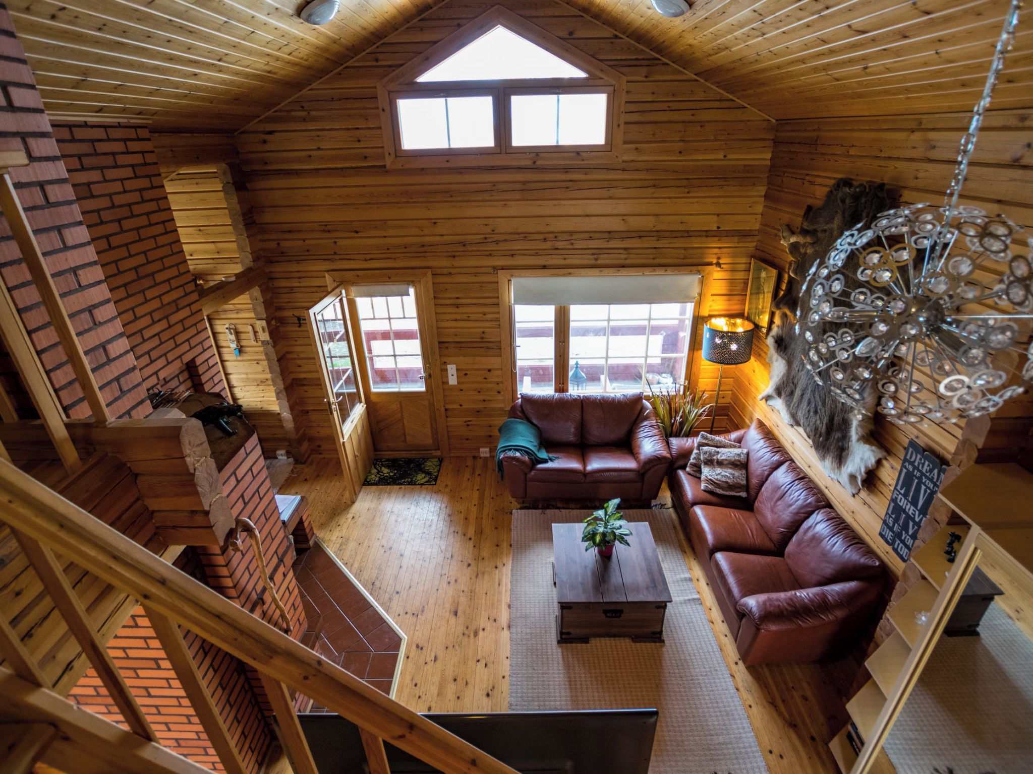 A wooden cabin interior with a loft bedroom, a living room with leather sofas, and a decorative metal chandelier.