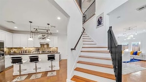 A modern kitchen with white cabinets, a central island with bar stools, and a staircase with wooden steps and black railings. - Luxurious 3BR 2.5BA Home with Hot Tub in Top Area (Houston)