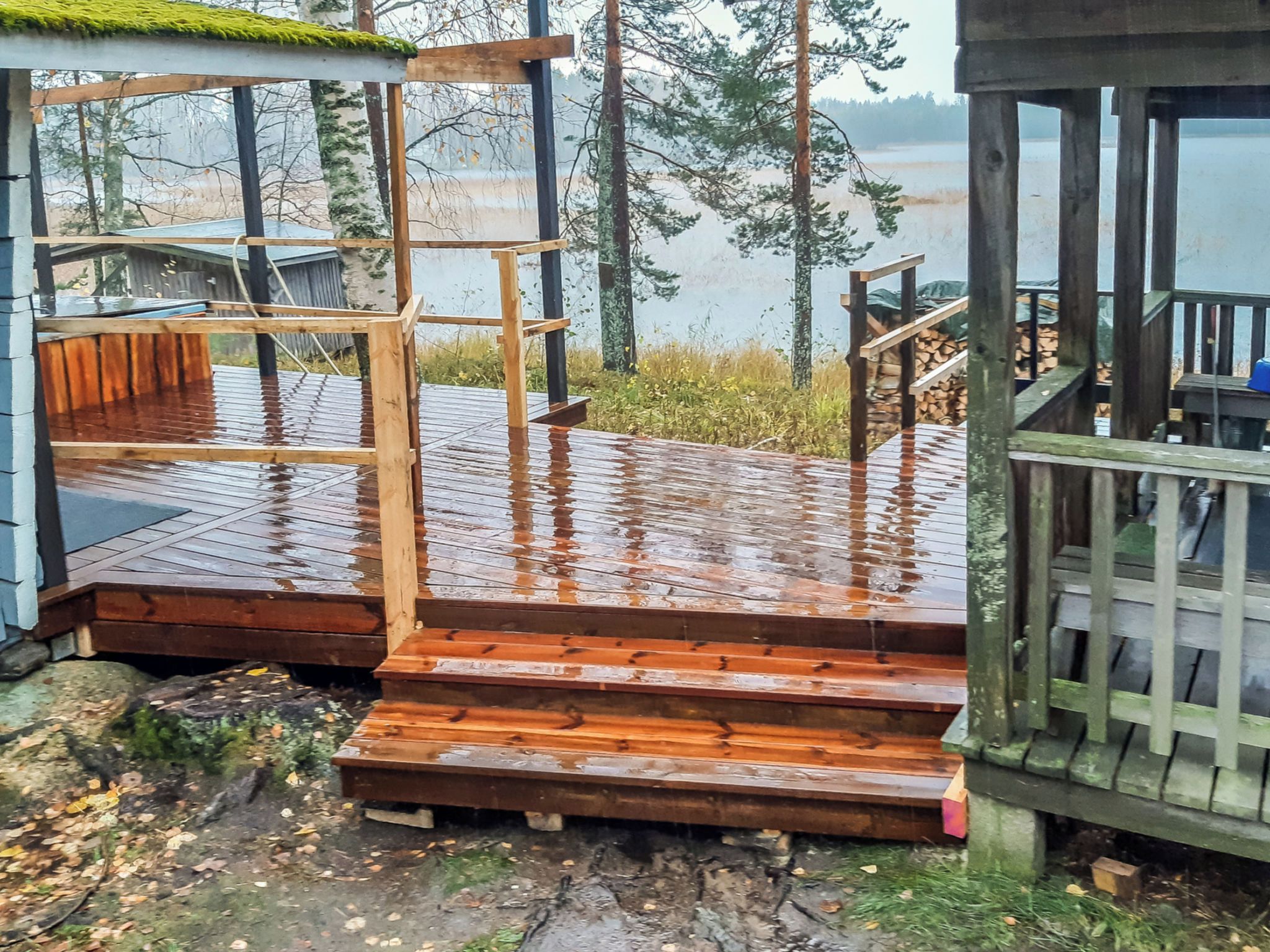 Una terraza de madera con escalones que conducen a una zona cubierta, rodeada de árboles y una masa de agua.