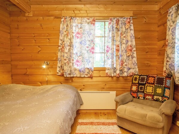 A cozy cabin room with a bed, a chair, a window with floral curtains, and a radiator.