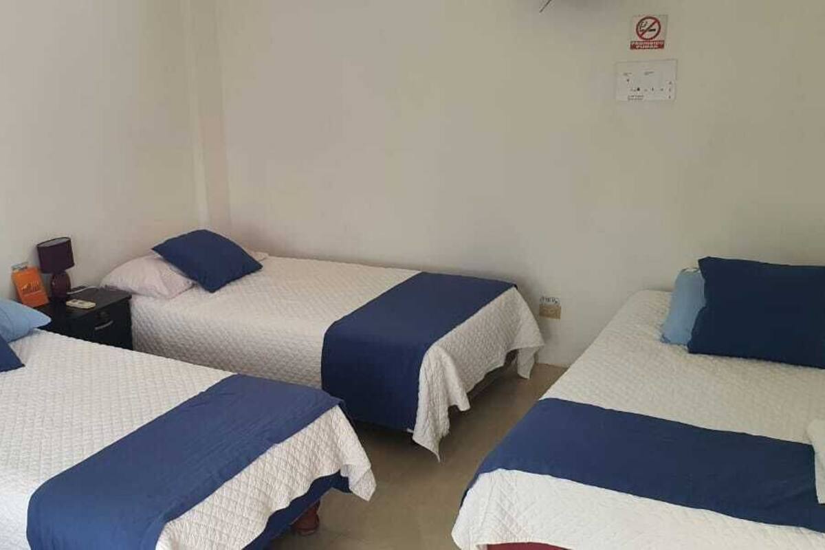 Une chambre avec trois lits simples, chacun recouvert d’une couvre-lit bleue et blanche, assortie aux oreillers. On y trouve une table de chevet avec une lampe et quelques objets.