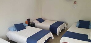 Ein Zimmer mit drei Einzelbetten, jedes mit einer blau-weißen Tagesdecke und passenden Kissen. Ein Nachttisch mit Lampe und einigen Gegenständen steht daneben.