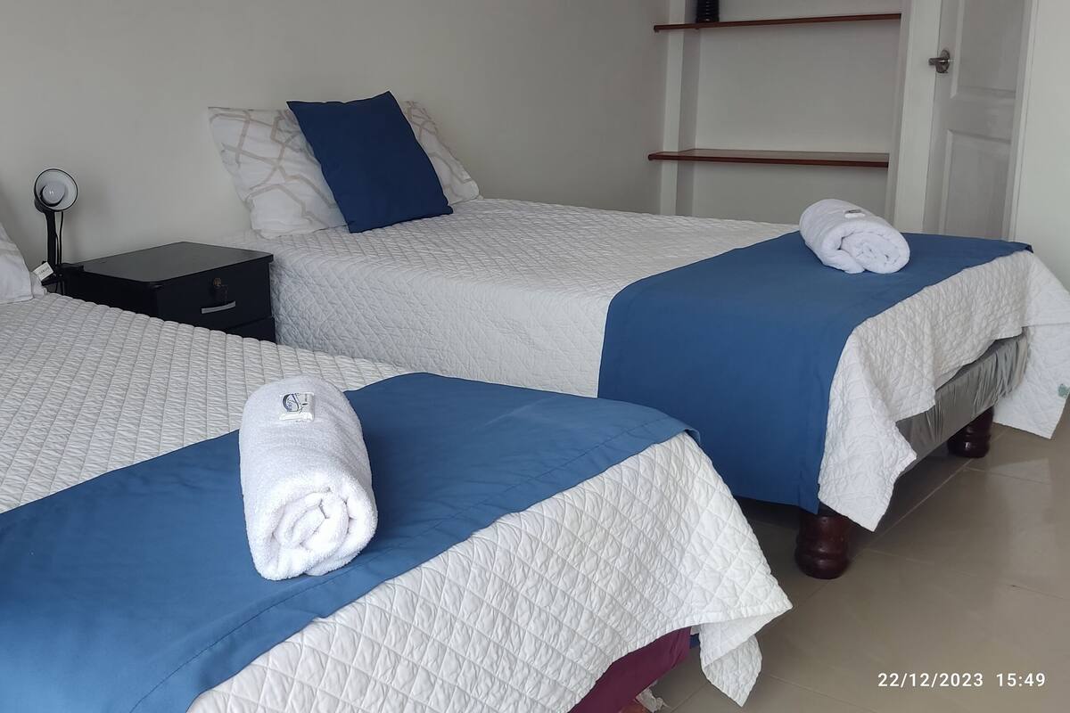 Une chambre d’hôtel avec deux lits, chacun recouvert d’une courtepointe matelassée bleue et blanche, une table de chevet avec une lampe, et une serviette roulée sur chaque lit.