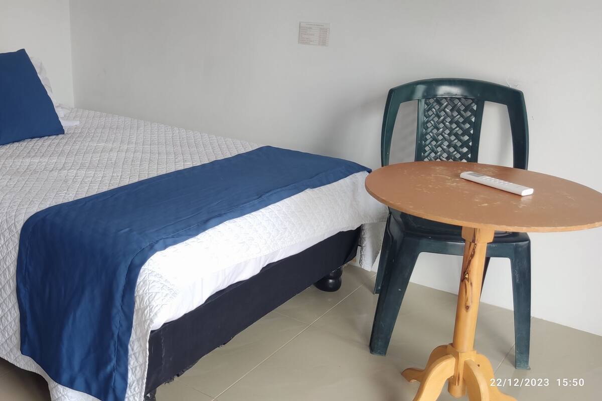 Un lit bien fait, recouvert d’une couette blanche et bleue, avec un oreiller bleu et une télécommande posée sur une petite table ronde à côté.