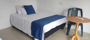 Ein ordentlich bezogenes Bett mit einer weiß-blauen Steude, einem blauen Kopfkissen und einer Fernbedienung auf einem kleinen runden Tisch daneben.