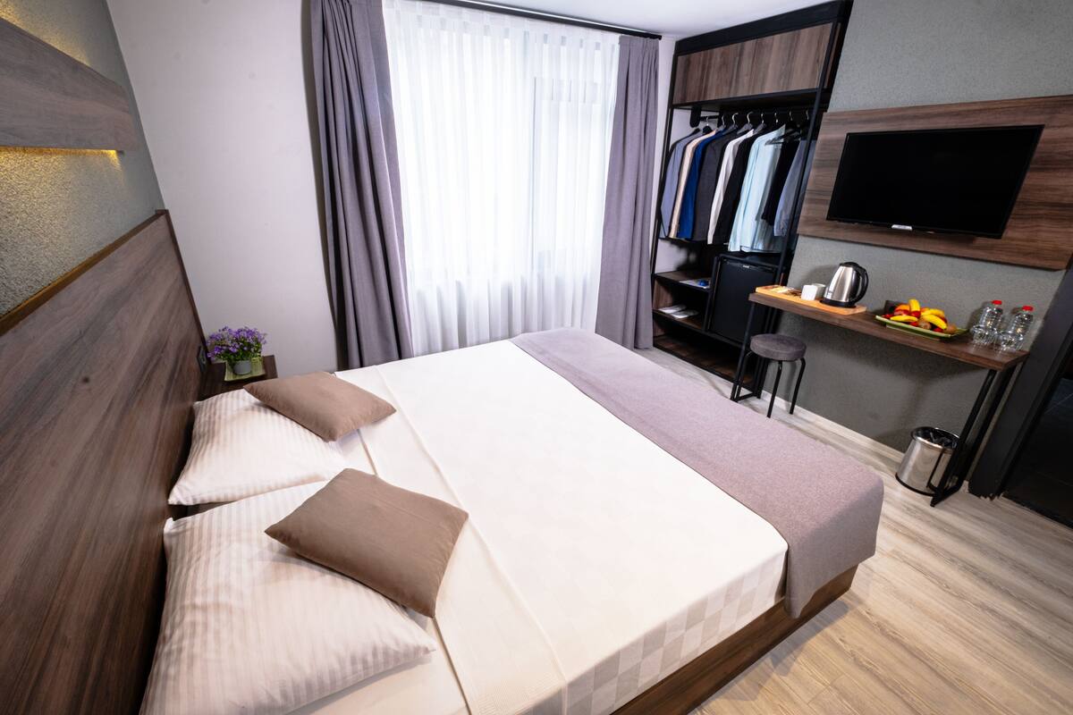 A modern hotel room with a large bed, a TV mounted on the wall, a wardrobe, and a small table with a kettle and fruit.