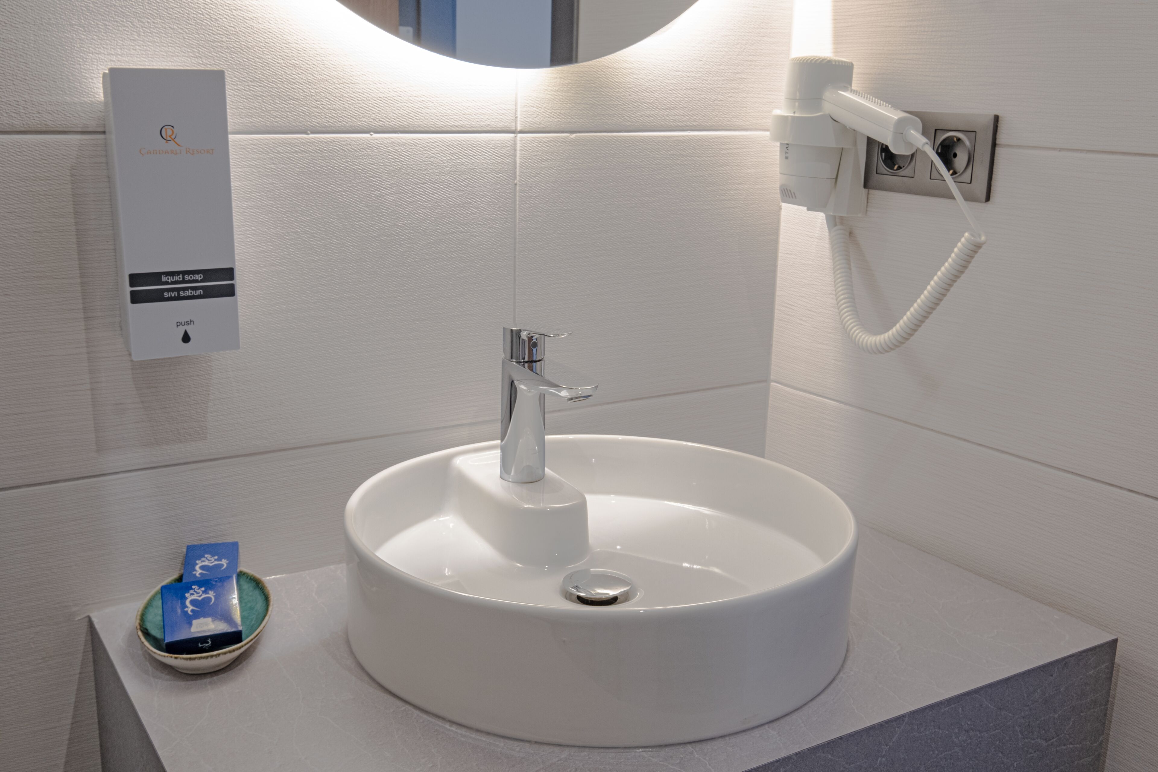 Een moderne badkamer met een witte wastafel, een spiegel en een zeepdispenser.