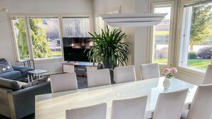 A modern living room with a glass dining table, white chairs, a grey sofa, a TV, and a large plant.
