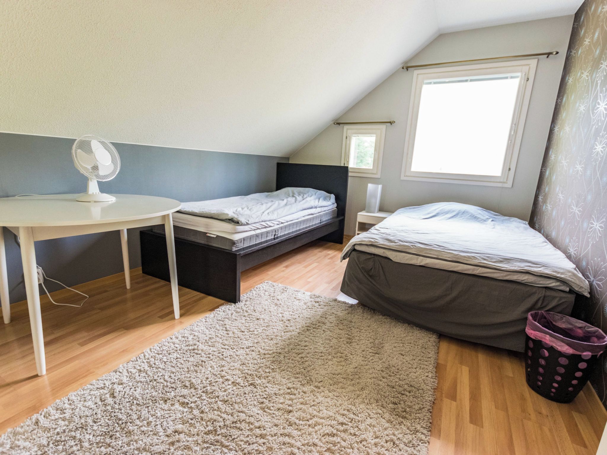A small, well-lit attic bedroom with a desk, two beds, a large window, and a ceiling fan.