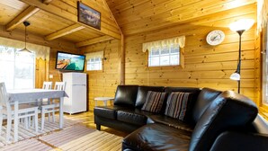 A cozy wooden cabin interior with a black leather sofa, a dining table with chairs, a small refrigerator, and a television mounted on the wall.