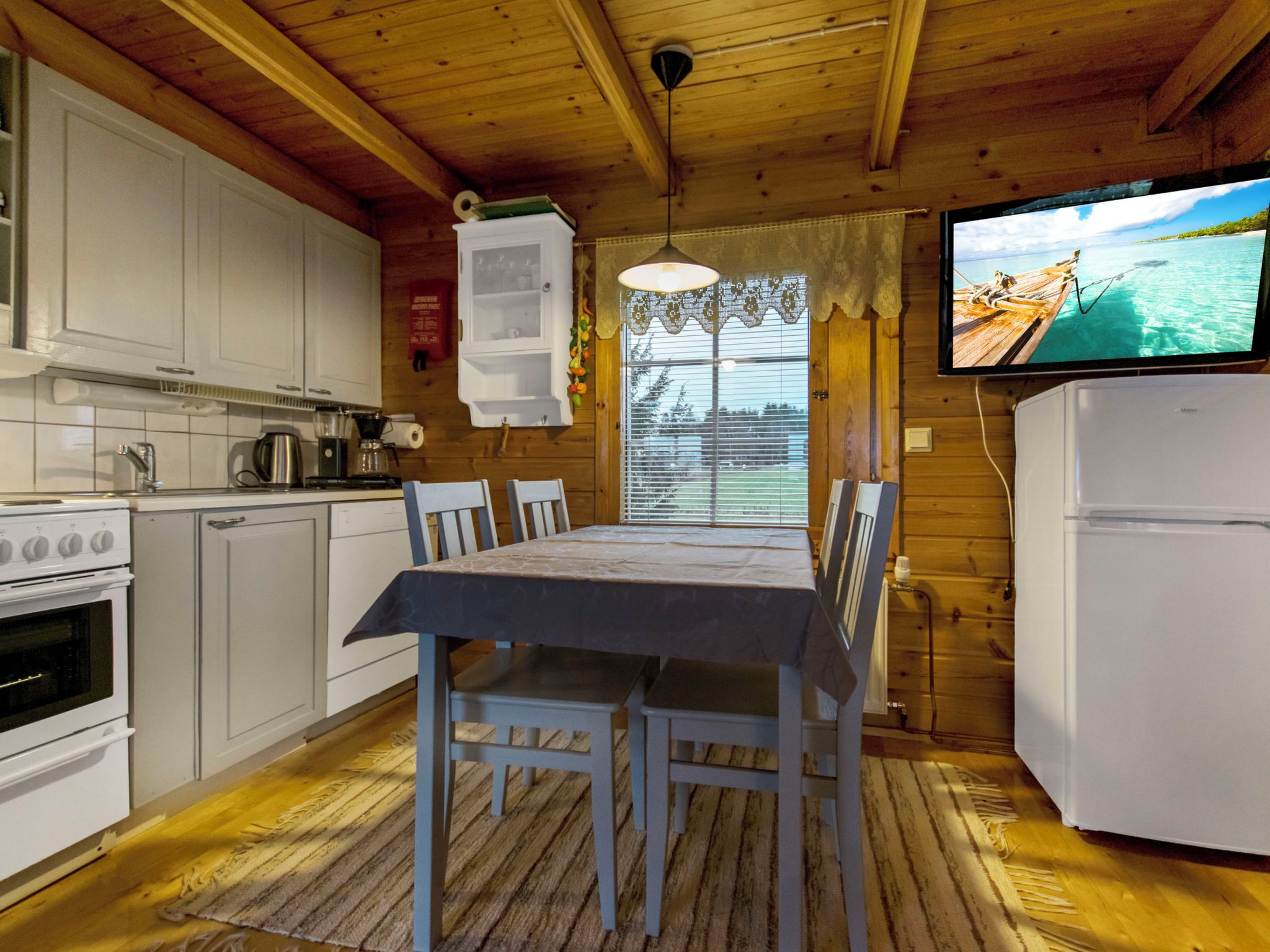 Ein Interieur einer Holzhütte mit einer Küche, einem Esstisch, Stühlen, einem Kühlschrank und einem Fernseher, der eine Strandansicht zeigt.