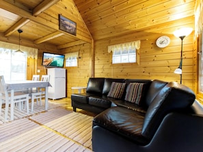 Ein Interieur einer Holzhütte mit einem schwarzen Ledersofa, einem weißen Esstisch mit Stühlen, einem kleinen weißen Kühlschrank und einem an der Wand befestigten Fernseher.