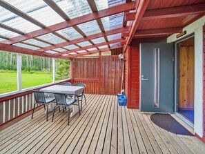 A covered outdoor deck with a table and chairs, a door, and a view of a green landscape.