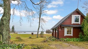 A red wooden cabin with a sloped roof, a small porch, and a window with shutters. A gravel path leads to the cabin, and there is a body of water in the background.
