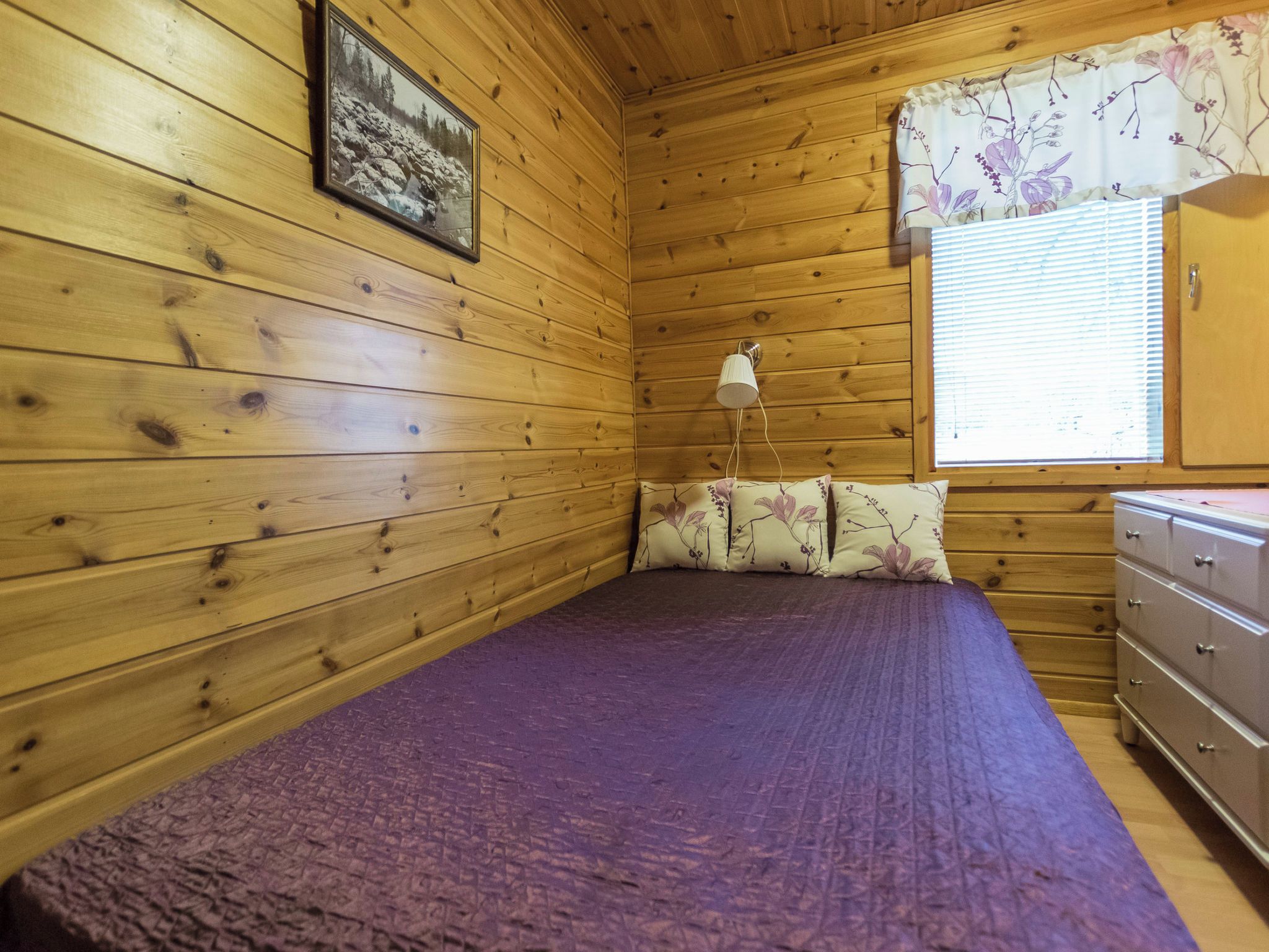 Ein Zimmer in einer Holzhütte mit einem Bett, einem Nachttisch, einer Lampe und einem Fenster mit Jalousien.