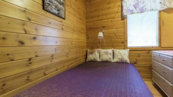 Ein Zimmer in einer Holzhütte mit einem Bett, einem Nachttisch, einer Lampe und einem Fenster mit Jalousien.