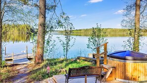 Una terraza de madera con un jacuzzi, un banco y una escalera de madera que conduce a un muelle junto a un lago tranquilo rodeado de árboles.