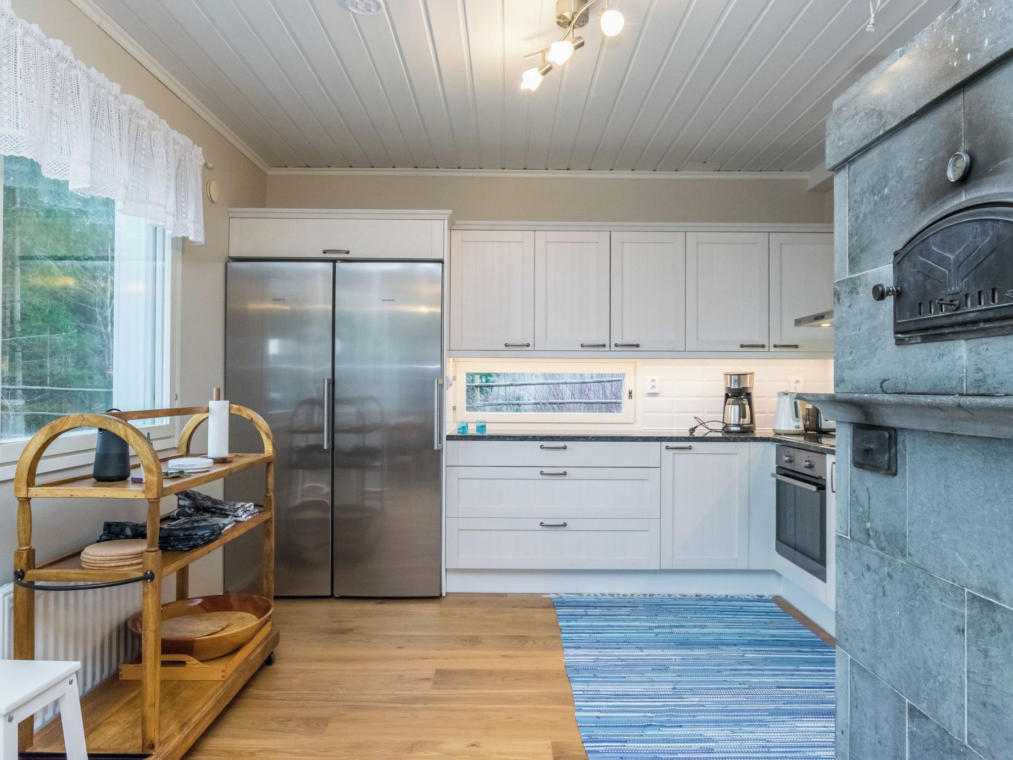 A modern kitchen with white cabinetry, a stainless steel refrigerator, a built-in oven, and a stone fireplace.