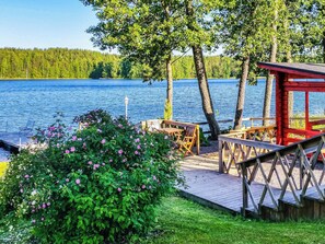 En træterrasse ved søen med træmøbler og en rød bygning, omgivet af grønt og træer.