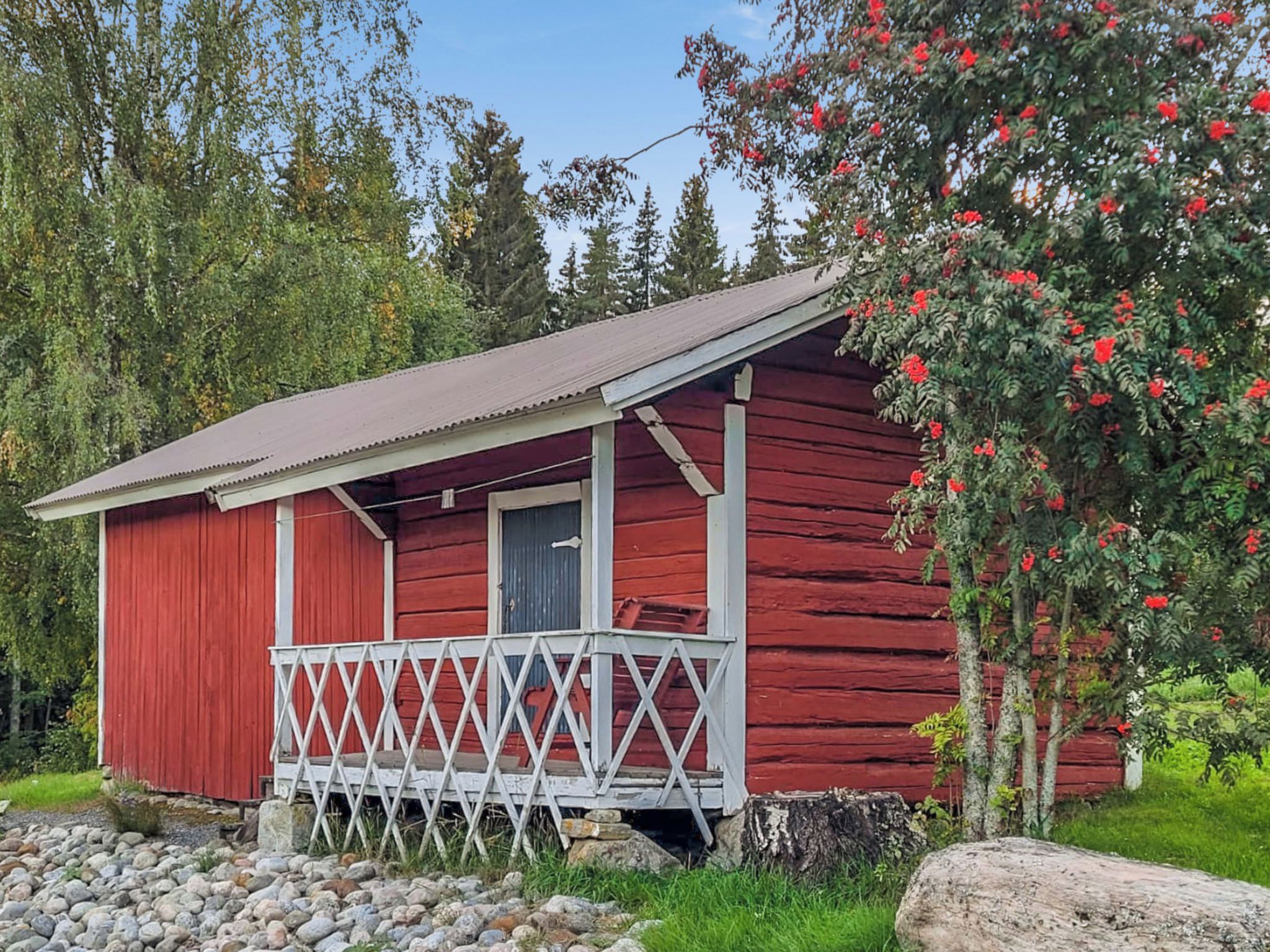 A red wooden cabin with a porch and a door, surrounded by greenery and trees.