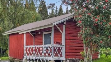 A red wooden cabin with a porch and a door, surrounded by greenery and trees.