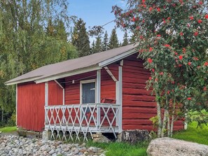 Una cabaña de madera roja con un porche y una puerta, rodeada de vegetación y árboles.